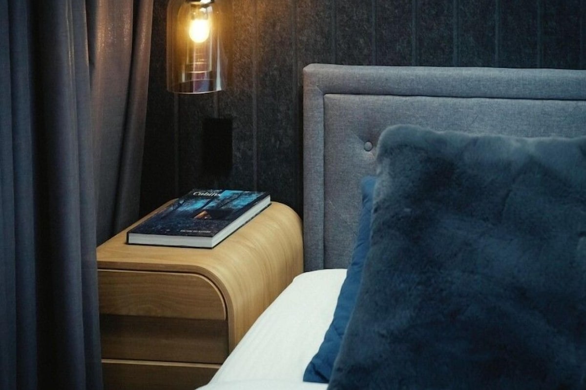 Una cama bien hecha con un cojín azul, una mesita de noche con un libro y una lámpara con pantalla transparente.