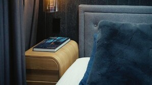 Una cama bien hecha con un cojín azul, una mesita de noche con un libro y una lámpara con pantalla transparente.