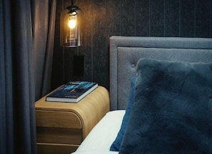 Un lit bien fait, avec un oreiller bleu, une table de chevet sur laquelle repose un livre et une lampe dotée d’un abat-jour transparent.