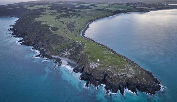 Una vista aerea di un paesaggio costiero con una stretta lingua di terra che conduce a una distesa d'acqua, circondata da campi verdi e terreno roccioso.