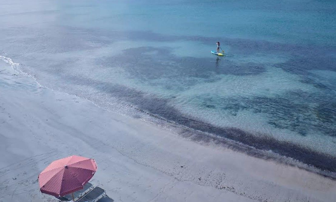 An aerial view of a beach with clear water, a person paddleboarding, and a red umbrella on the sand.