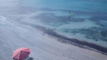 An aerial view of a beach with clear water, a person paddleboarding, and a red umbrella on the sand.
