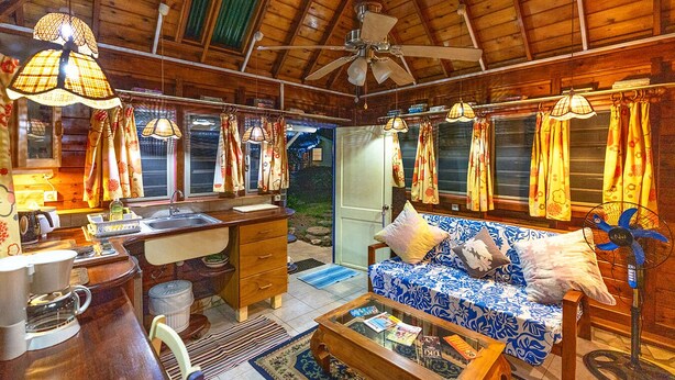 A cozy cabin interior with a wooden ceiling, a sofa with patterned cushions, a dining area with a table and chairs, and a window with curtains. - Fare Miti (Moorea-Maiao)
