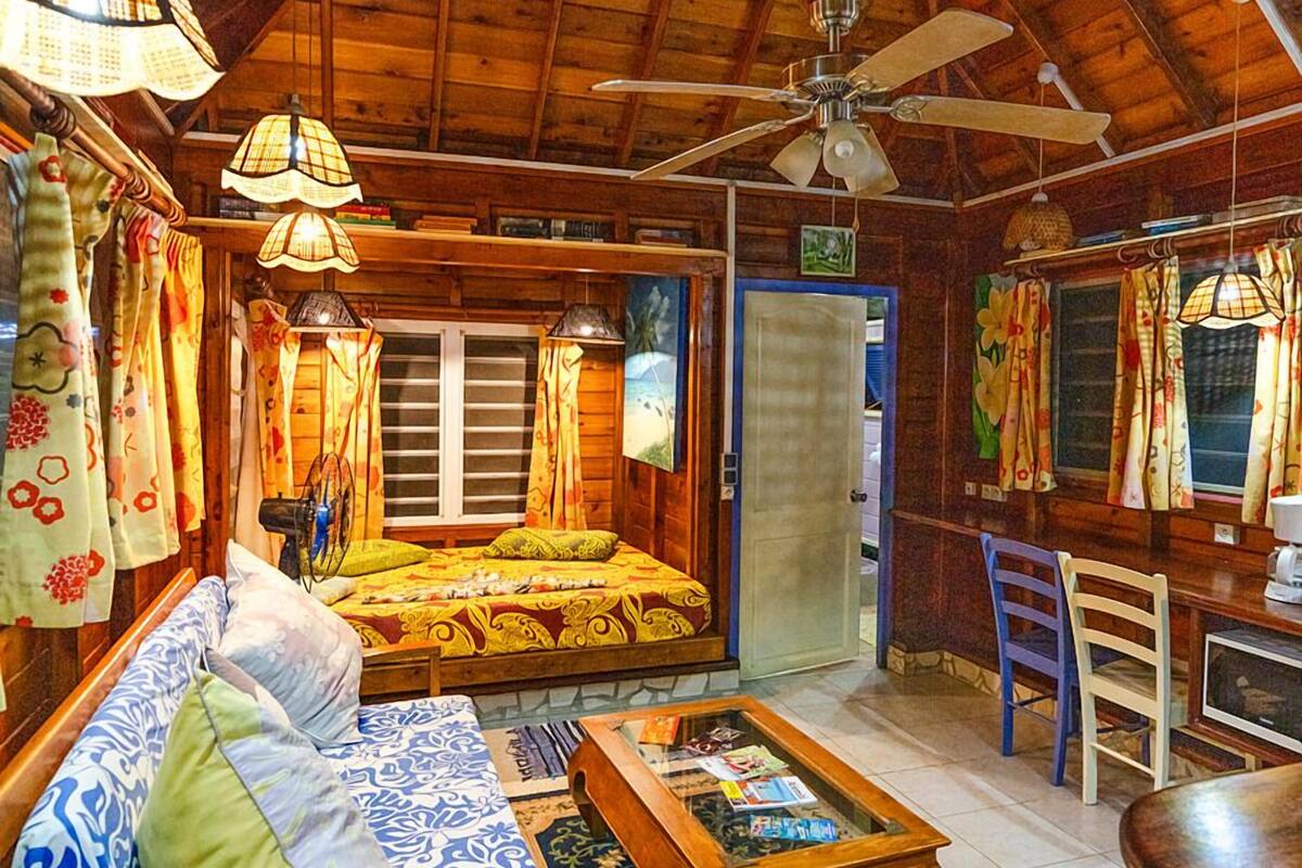 A cozy room with a bed, sofa, and a coffee table. There is a ceiling fan, a window with curtains, and a door.