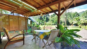 A covered outdoor seating area with wooden furniture and a table, surrounded by lush greenery and a view of a mountainous landscape. - Fare Miti (Moorea-Maiao)