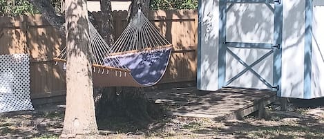 Relax in a serene hammock suspended between trees, surrounded by a garden with a small storage shed.