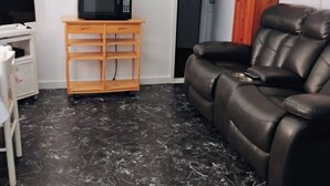 A living room with a black leather sofa, a wooden TV stand with a microwave, a marble floor, and artwork on the walls. - Charming 1-bedroom cottage in Port Orange Close to Daytona Beach  (Port Orange)