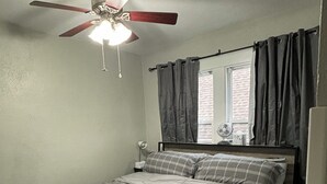 Un dormitorio con cama, ventilador de techo, ventana con cortinas y mesita de noche con lámpara.