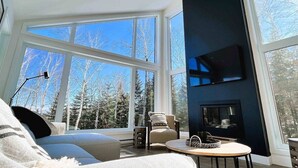 A modern living room with large windows, a flat-screen TV, and a cozy seating area. - Le Verseau 148: Hot Tub, Forest Views & BBQ (Baie-Saint-Paul)