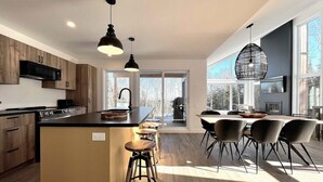 A modern kitchen with a central island, bar stools, and a dining area with chairs. - Le Verseau 148: Hot Tub, Forest Views & BBQ (Baie-Saint-Paul)