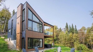 A modern two-story house with a large glass balcony, surrounded by a forested area. - Le Verseau 148: Hot Tub, Forest Views & BBQ (Baie-Saint-Paul)