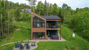 A modern two-story house with large windows, a deck, and outdoor seating. - Le Verseau 148: Hot Tub, Forest Views & BBQ (Baie-Saint-Paul)