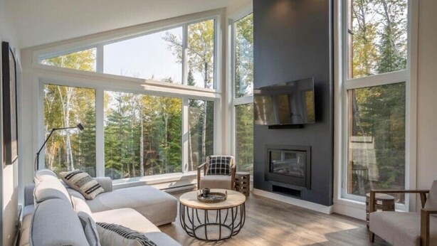 A spacious living room with a large sofa, a round coffee table, and a fireplace. - Le Verseau 148: Hot Tub, Forest Views & BBQ (Baie-Saint-Paul)