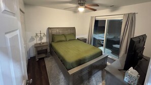 A bedroom with a bed, a ceiling fan, a nightstand, a lamp, a window with curtains, and a sliding door.