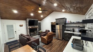 A modern kitchen with a built-in refrigerator, a dining area with a wooden table and chairs, and a living room with leather sofas and a stone fireplace.