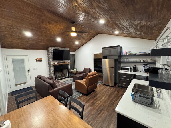 Una cocina moderna con refrigerador integrado, un comedor con mesa y sillas de madera, y una sala de estar con sofás de cuero y chimenea de piedra.