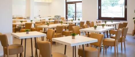 A spacious dining area with multiple tables and chairs, large windows, and indoor plants.