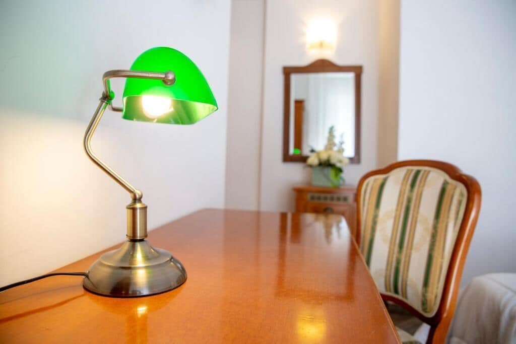 En lampe med grøn skærm, et træbord, en stol med stribet pude og et spejl på væggen.