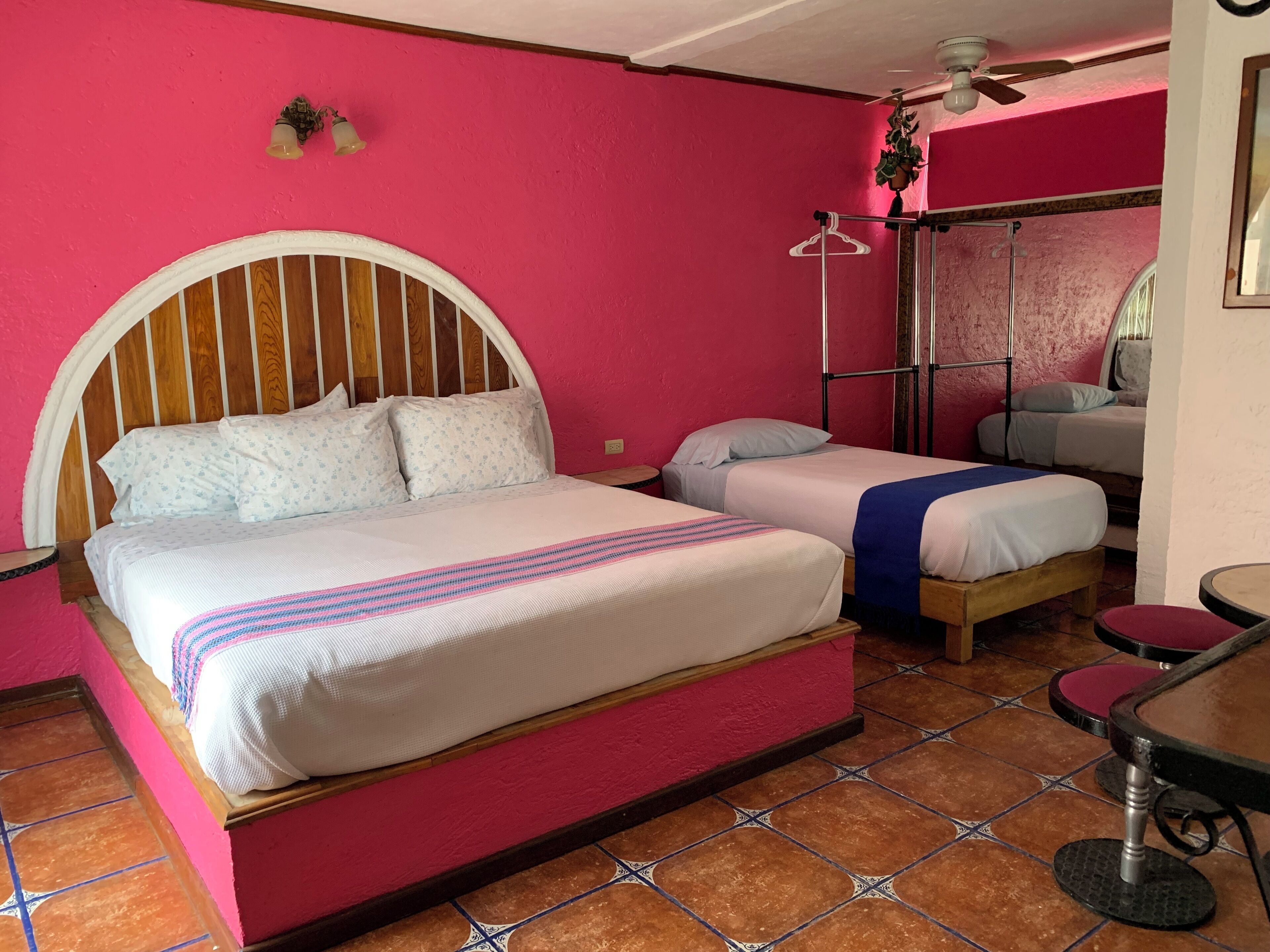 A room with a large bed, two smaller beds, a ceiling fan, a hanging plant, a mirror, and a tiled floor.