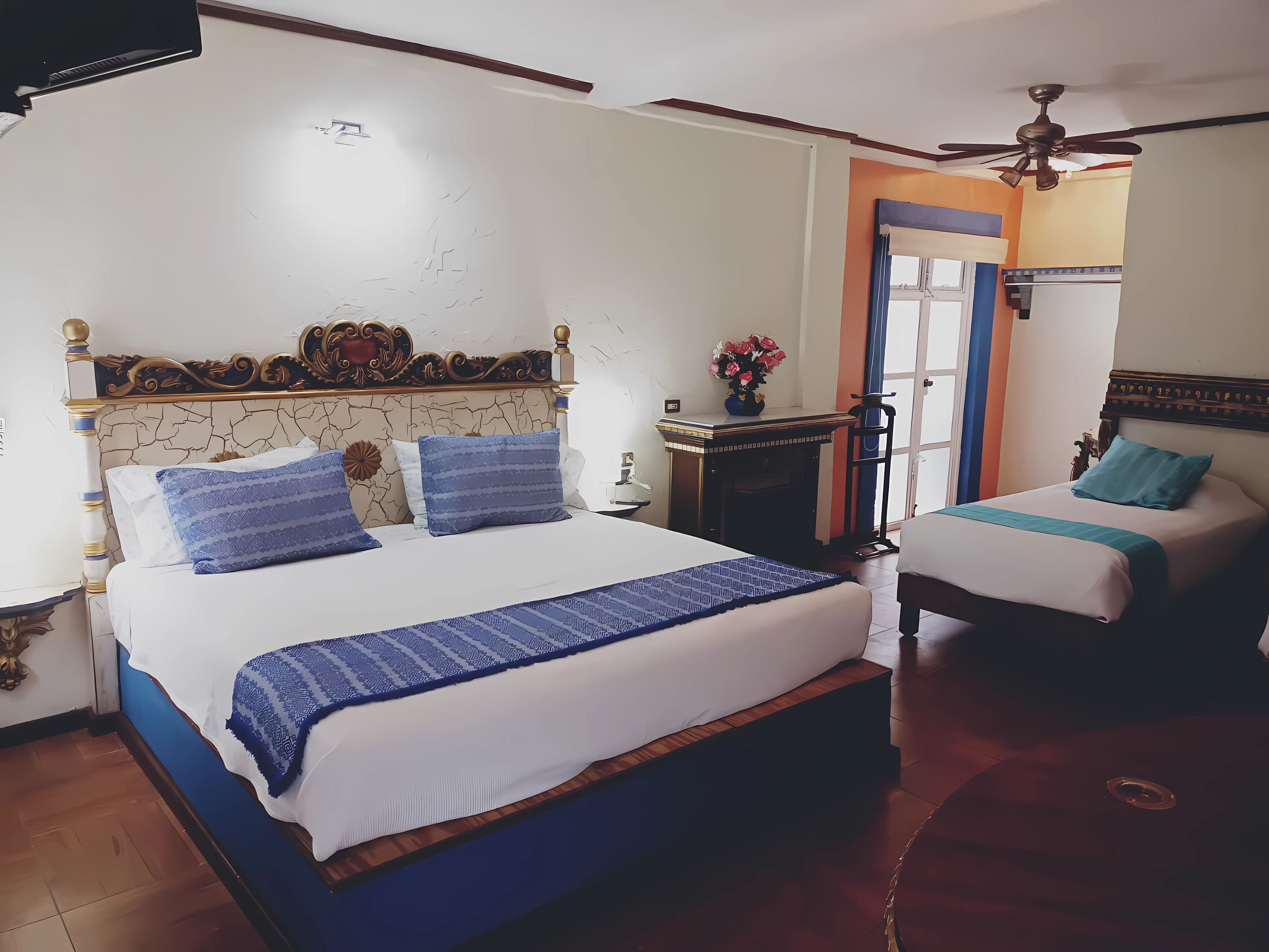 Une chambre d’hôtel avec deux lits, une petite table ornée d’un vase de fleurs, un ventilateur de plafond et une vue sur l’extérieur.
