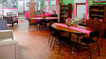 A dining area with a wooden table, black chairs, and a pink tablecloth. There are colorful vases on the table and a bookshelf with various items in the background.