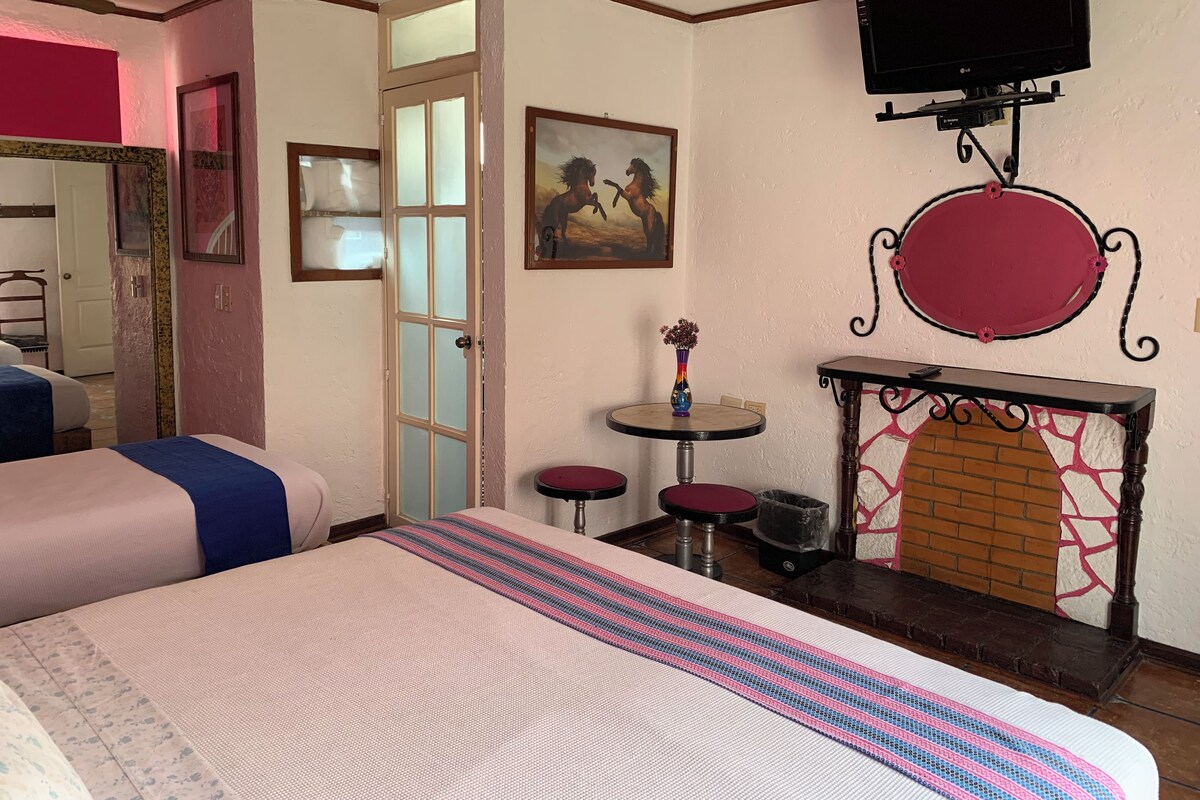 A hotel room with a bed, a small table with a vase, a fireplace, and a TV mounted on the wall.