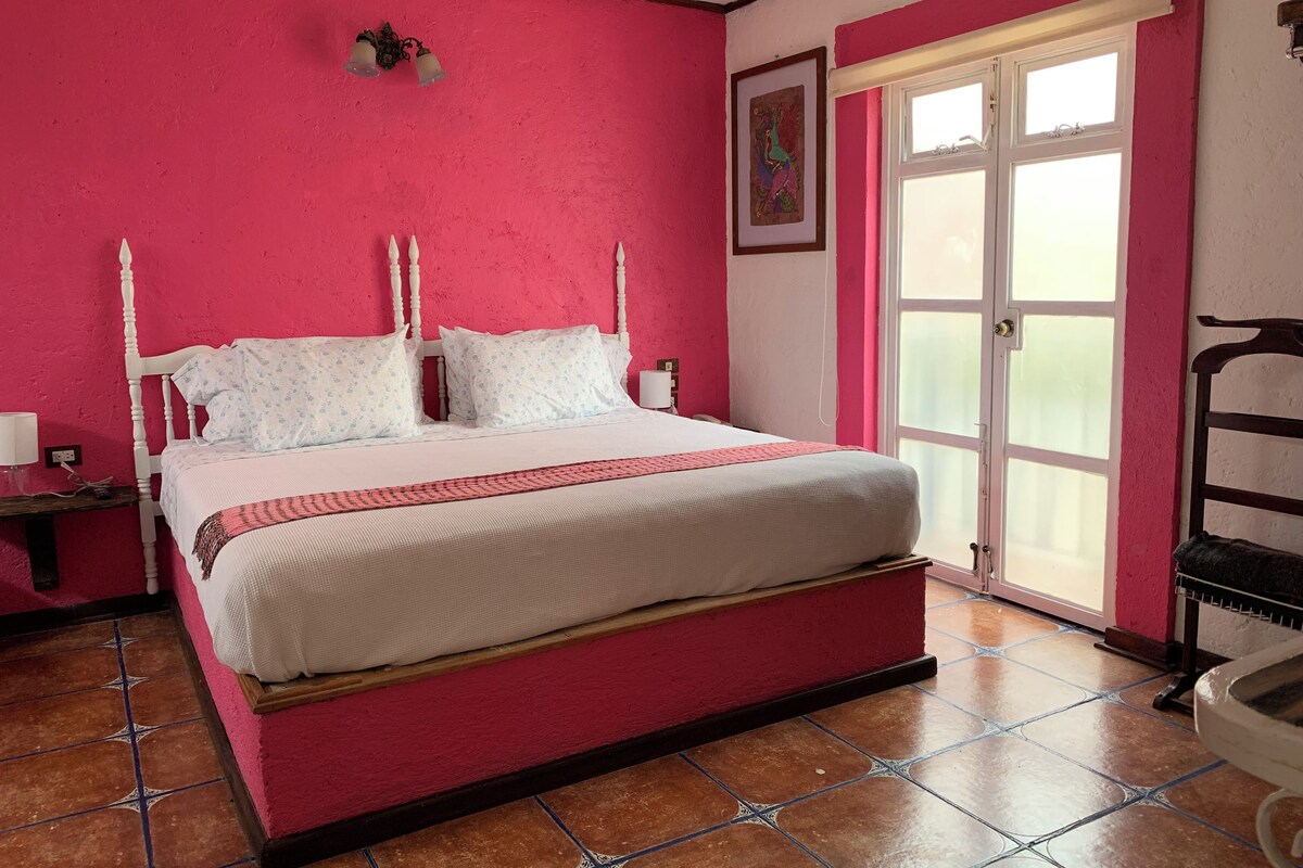 A bedroom with a large bed, a wooden nightstand, a wall-mounted lamp, and a window with a view.