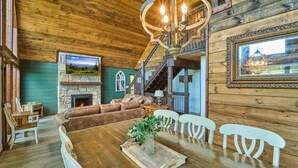 A rustic wooden interior with a dining table, white chairs, a fireplace, and a staircase leading to an upper level. - Family Friendly Getaway I Hot Tub In Gatlinburg (Gatlinburg)