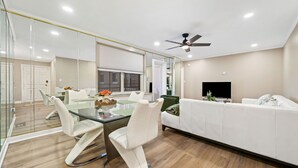 A modern living room with a glass dining table, white sofas, and a ceiling fan.