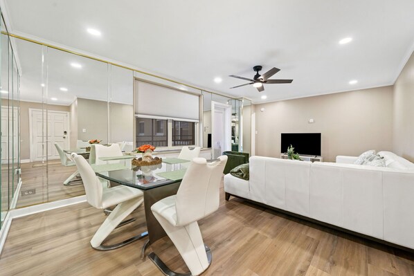 A modern living room with a glass dining table, white sofas, and a ceiling fan.