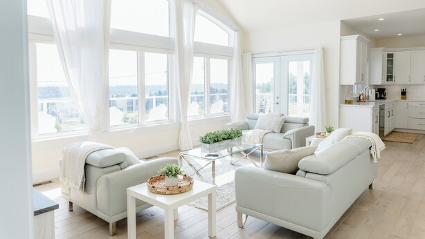 A spacious living room with a large window, a sofa, a coffee table, and a kitchen area visible through an open door.