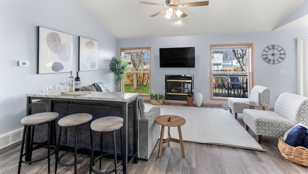 Un salon moderne comprenant un coin cuisine, un ventilateur de plafond, une cheminée et une grande fenêtre donnant sur un jardin.