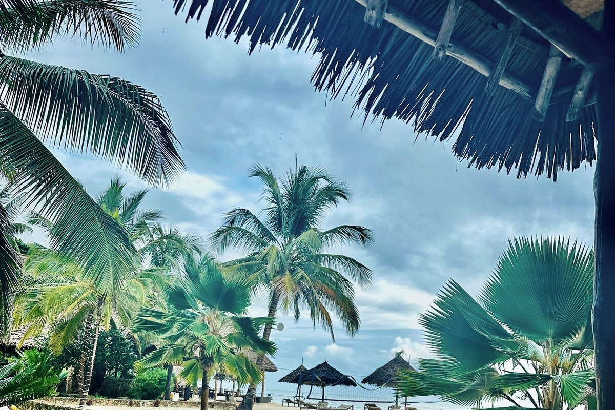 A view from a covered area with a palm tree and beach in the background.