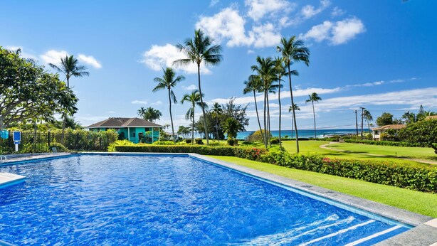 Ein Swimmingpool mit klarem, blauem Wasser, umgeben von gepflegter Bepflanzung und Palmen, mit Blick auf den Ozean und einem Gebäude im Hintergrund.