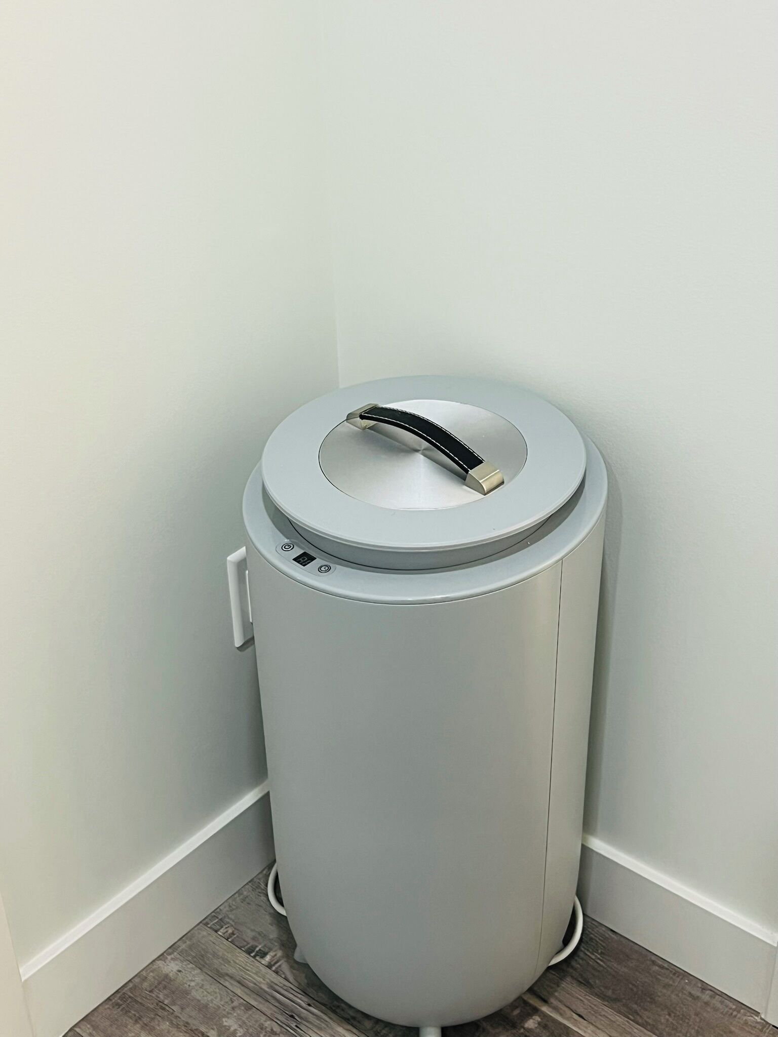 A compact, cylindrical trash bin with a lid and a handle, placed against a white wall with a wooden floor.