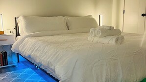A neatly made bed with white linens and towels, a bedside table with a lamp, and a window with blinds.