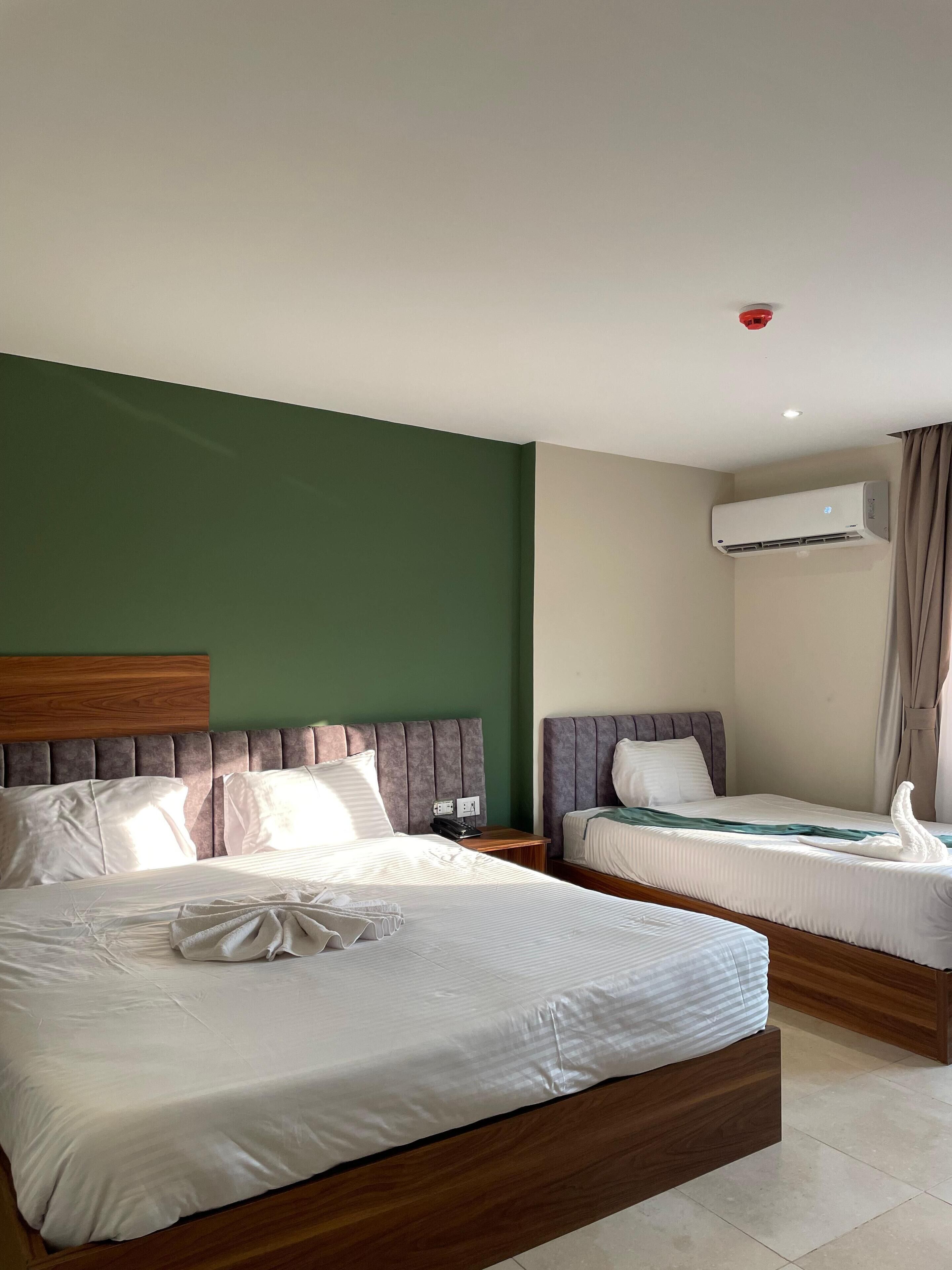 Une chambre d’hôtel avec deux lits, une tête de lit en bois, un mur vert, un appareil de climatisation et un luminaire rouge au plafond.