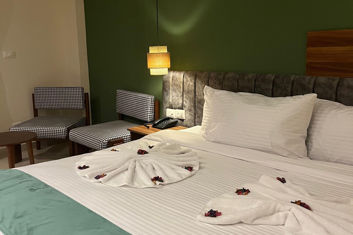 A hotel room with a bed, a bedside table, a chair, an air conditioning unit, and a wall-mounted lamp.