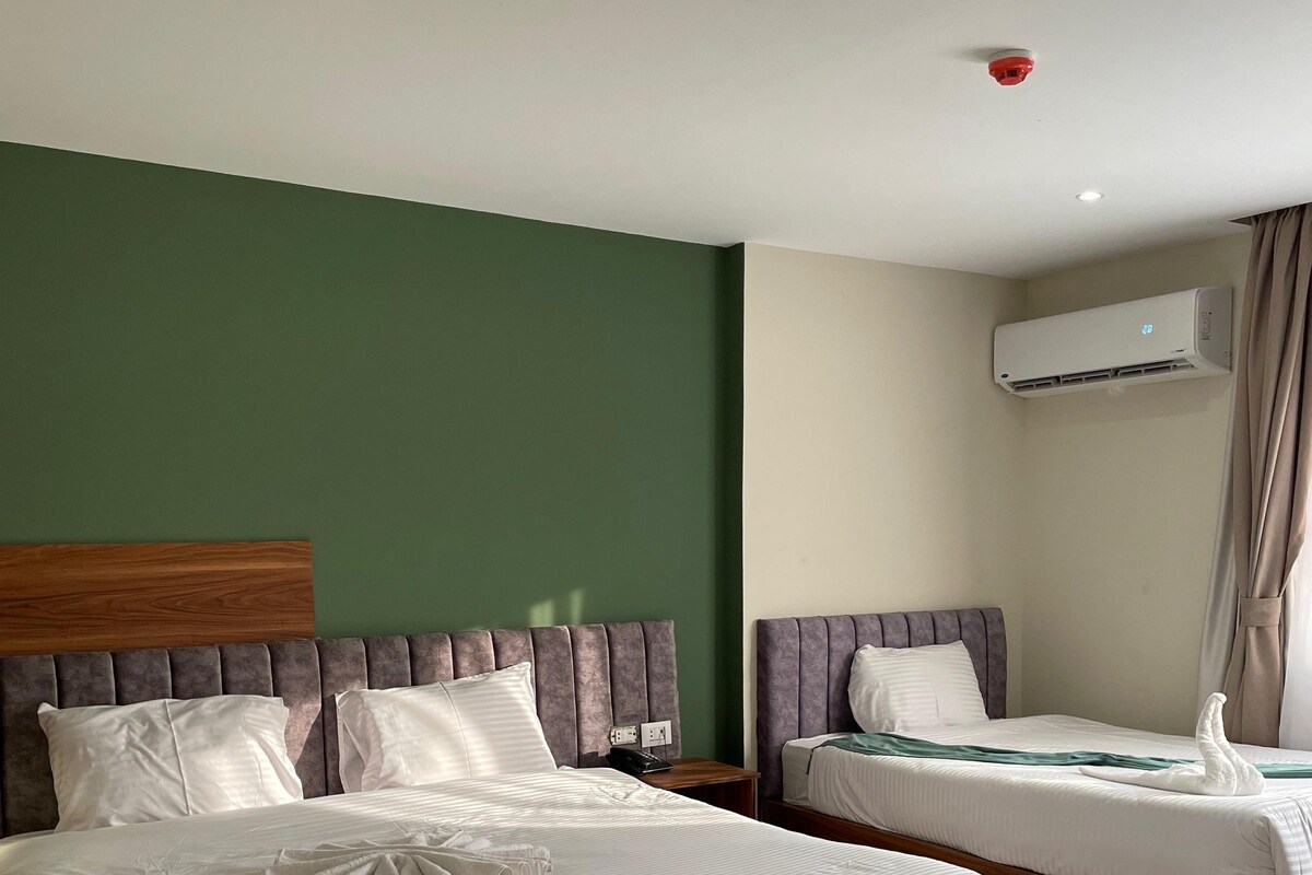Une chambre d’hôtel avec deux lits, un mur vert, un appareil de climatisation et un détecteur de fumée fixé au plafond.