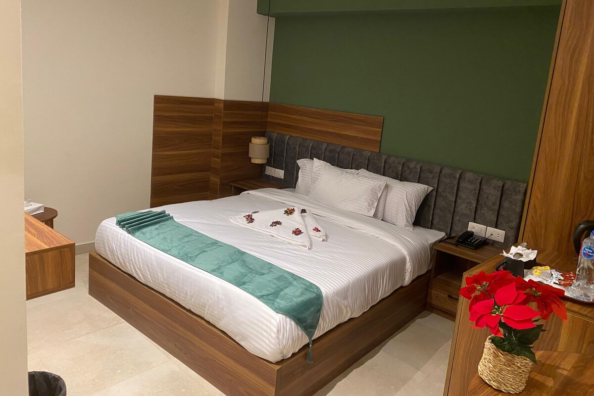 A neatly made bed with white linens and a teal throw, a bedside table with a lamp, and a nightstand with a phone and a bottle.