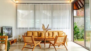 Un salon moderne avec un canapé en bois, deux fauteuils en osier, une table basse en verre et un téléviseur posé sur un meuble en bois.