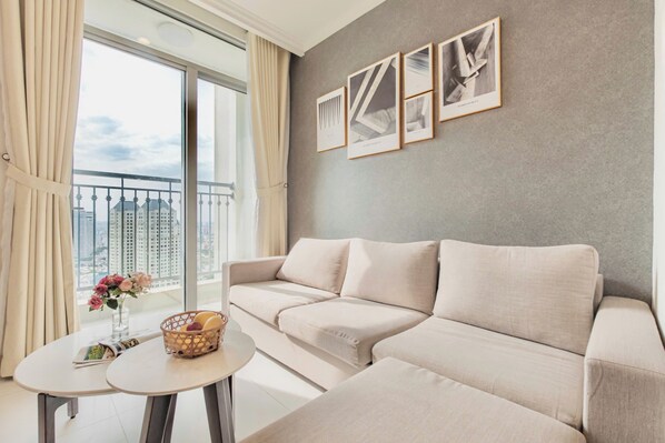 Una sala moderna con un sofá color beige, una mesita redonda con un jarrón de flores y vistas de la ciudad a través de amplias ventanas.