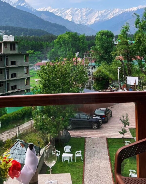 A balcony with a view of a mountainous landscape, a parked car, and a table with chairs and a wine glass. - Snow Park Villas & Resort Manali (Manali)