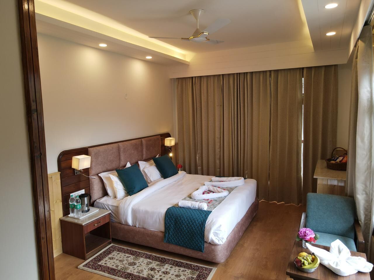 A hotel room with a large bed, a nightstand, a chair, a table with a bowl of fruit, and a window with curtains.