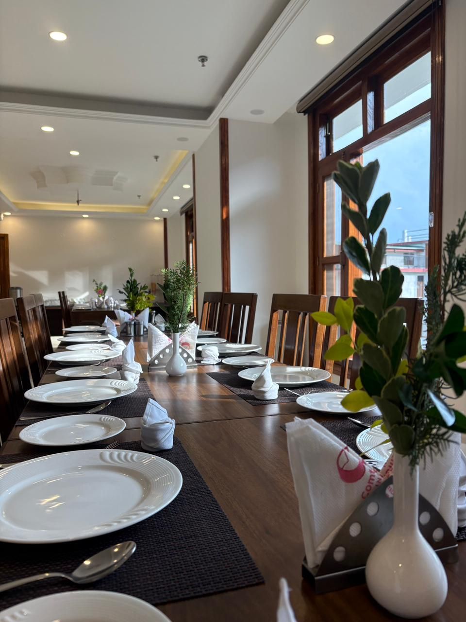 Un espace repas avec une longue table dressée pour un repas, entourée de chaises, de grandes fenêtres et agrémenté de plantes d’intérieur.