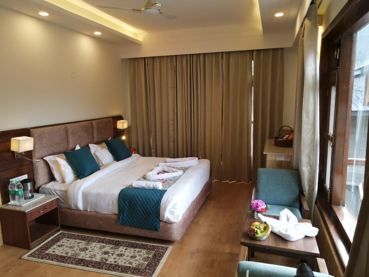 A hotel room with a large bed, a bedside table with a lamp, a small table with a fruit bowl and a vase, and a window with curtains.