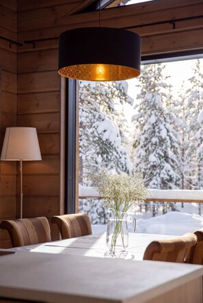 Un intérieur en bois avec une table à manger, des chaises, une lampe et une vue sur les arbres enneigés à l’extérieur.
