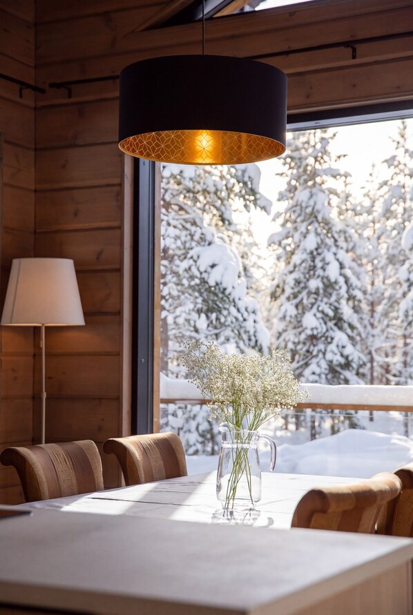 A wooden interior with a dining table, chairs, a lamp, and a view of snow-covered trees outside.
