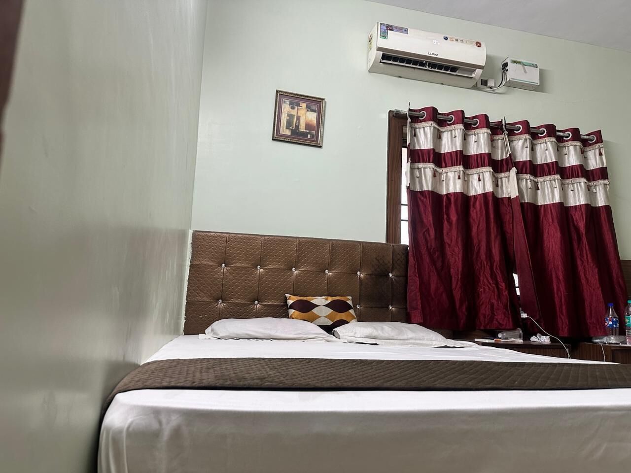 Une chambre d’hôtel avec un lit, une tête de lit, un appareil de climatisation, un tableau encadré au mur et des rideaux bordeaux.
