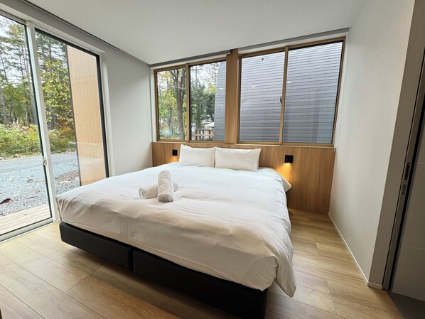 A modern bedroom with a large bed, wooden headboard, and a view of trees through the windows. - Everglow Hakuba (Hakuba)